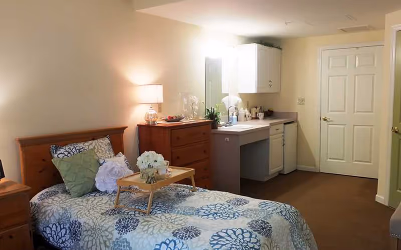 A cozy bedroom with a single bed covered in a floral patterned bedspread and several pillows. A wooden nightstand with a lamp is beside the bed. Next to the bed is a wooden dresser with a lamp and decorative items on top. In the background, there is a small vanity area with a sink, mirror, and cabinets. The room has beige walls and carpeted floor.