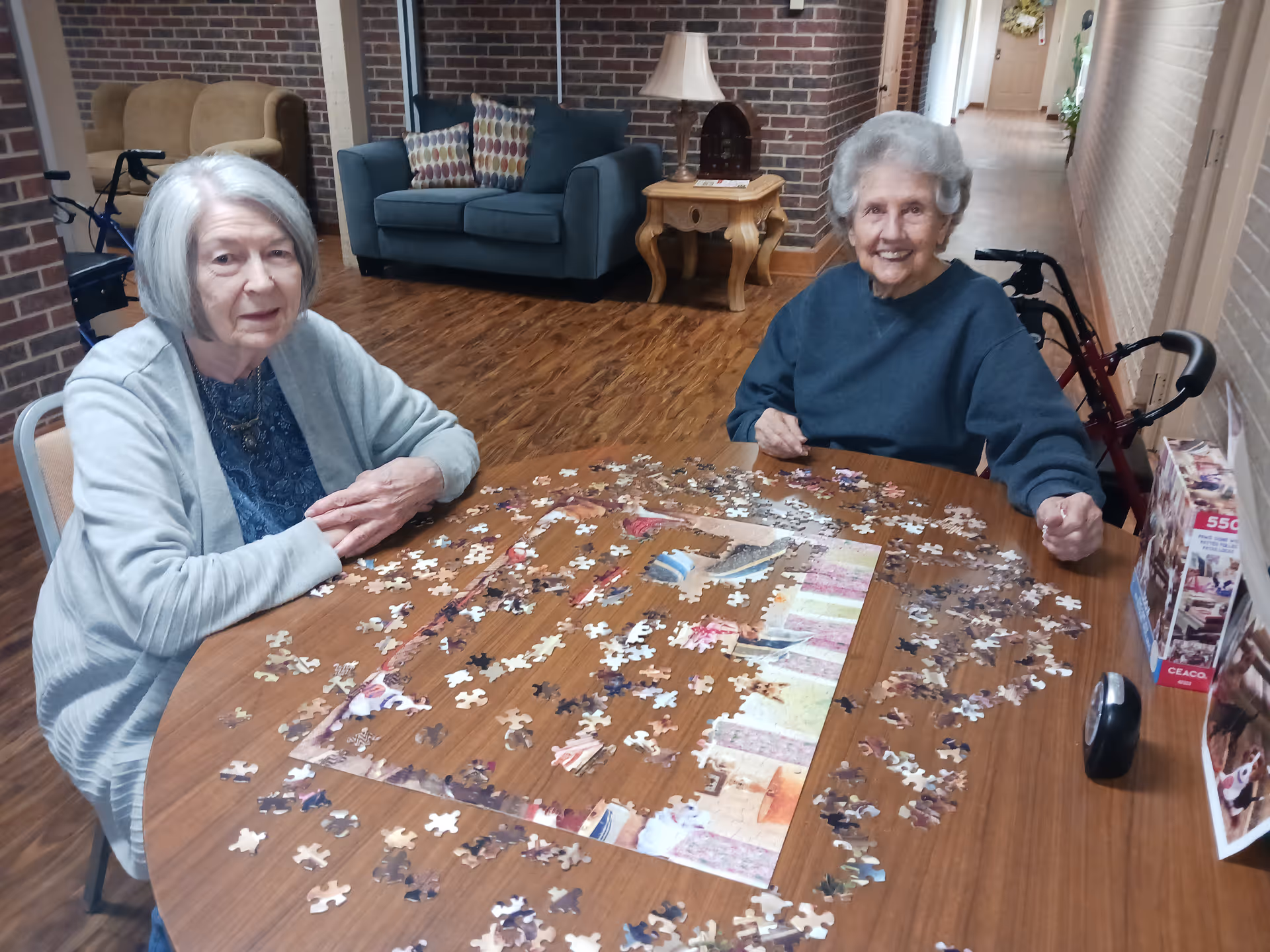 Two elderly women sitting at a wooden table working on a partially completed jigsaw puzzle in a cozy room with wooden flooring, a blue couch, a side table with a lamp, and brick walls. One woman has gray hair and is wearing a light gray cardigan, while the other has white hair and is wearing a dark blue sweater. A walker is visible behind each woman.