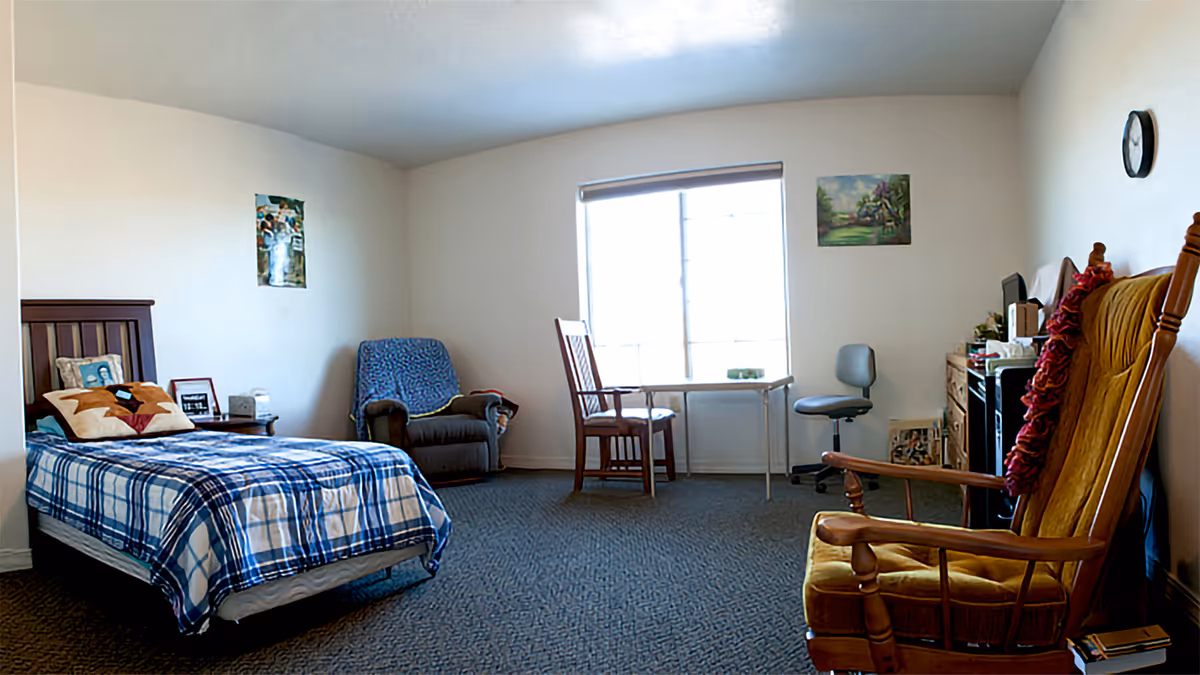 A simple bedroom in an assisted living facility with a single bed covered in a blue and white plaid blanket, a wooden headboard, and pillows. There is a small nightstand with framed photos and a lamp next to the bed. A cushioned armchair with a blue cover is placed in the corner. A window with a roller shade lets in natural light, and in front of it is a small table with two chairs, one wooden and one office-style. On the right side, there is a wooden rocking chair with a red cushion, a clock on the wall, and a dresser with various items on top. The walls are plain white with a couple of small paintings.