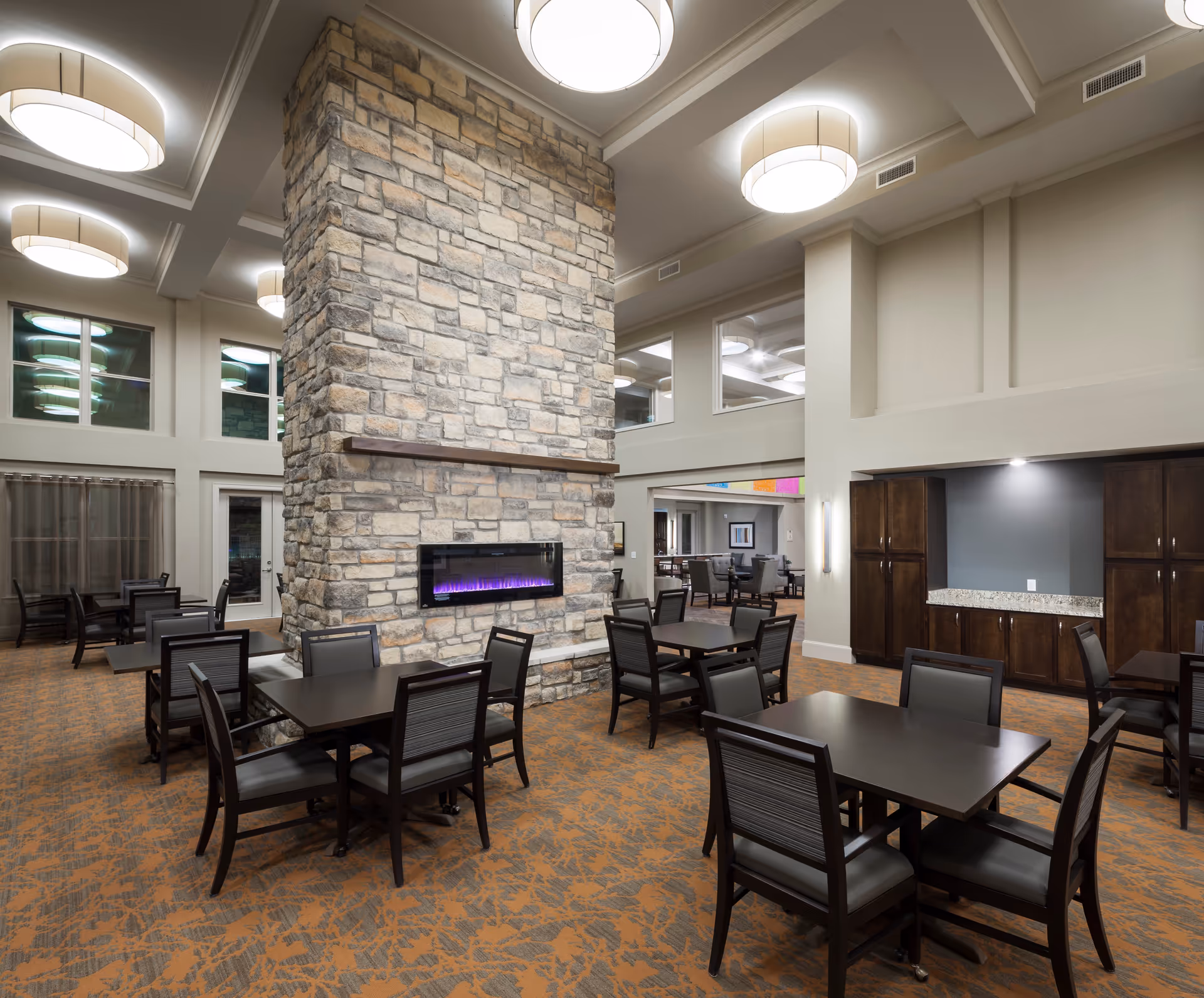 Spacious dining and common area with multiple tables and chairs centered around a tall stone fireplace and modern ceiling lights.