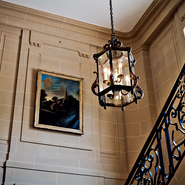 Interior view of a stairwell with beige stone walls, a decorative black wrought iron railing, a hanging lantern-style chandelier with lit bulbs, and a framed painting depicting a landscape with trees and a tall spire.