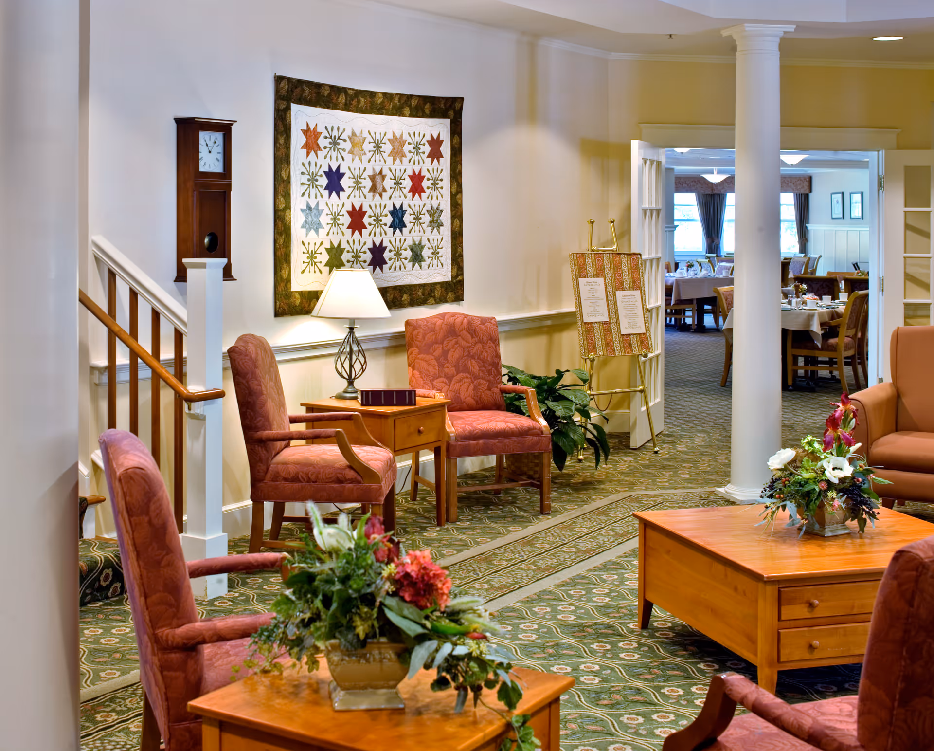 A cozy sitting area in a senior living facility with upholstered armchairs arranged around wooden tables adorned with floral arrangements. A quilt with star patterns hangs on the wall above a table lamp, and a wooden grandfather clock is mounted near a staircase. In the background, an open doorway leads to a dining room with tables and chairs set for a meal.