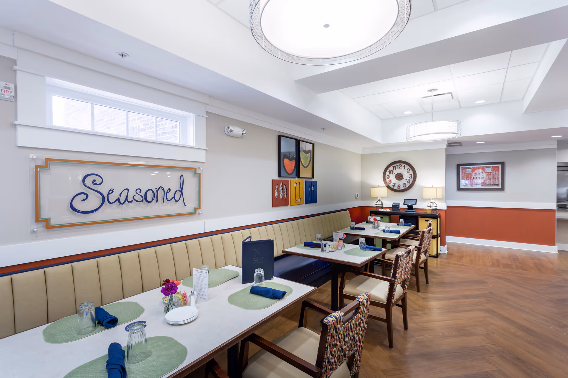 A dining area with tables set with placemats, napkins, glasses, and plates. There is a long cushioned bench along the wall with several chairs on the opposite side of the tables. The wall features a sign that says 'Seasoned' and colorful artwork. The room has wood flooring, modern light fixtures, and a large clock on the far wall.