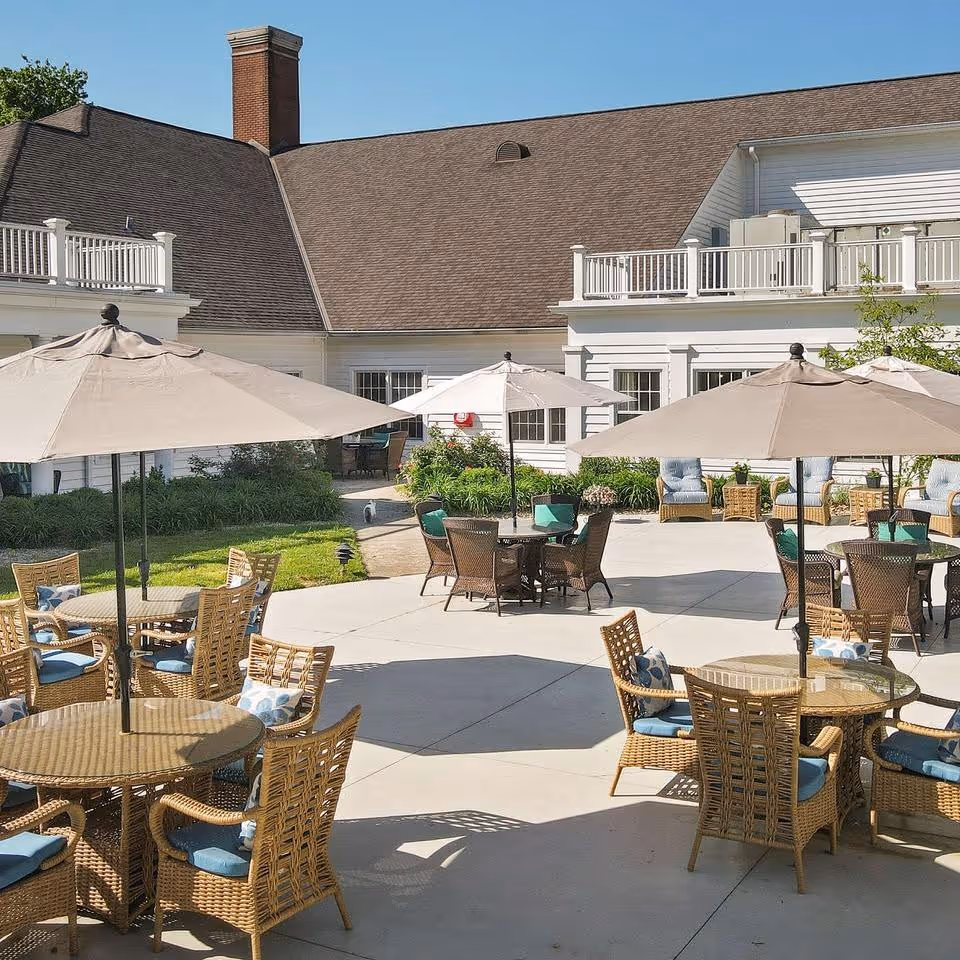 Outdoor patio area with multiple round tables and wicker chairs, each table shaded by large umbrellas. The patio is adjacent to a white building with a brown roof and a balcony. There are some plants and greenery around the patio area under a clear blue sky.