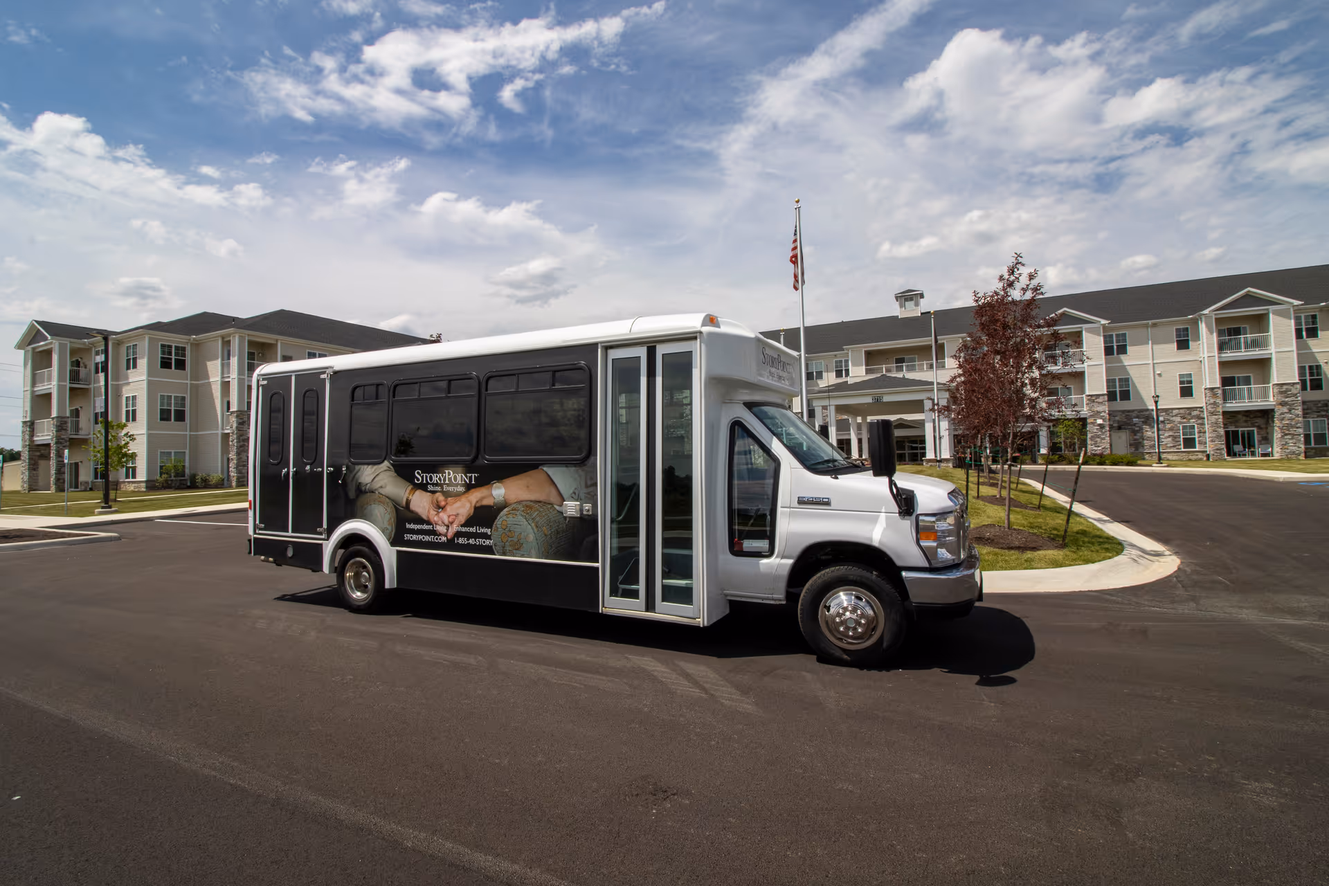 A white shuttle bus with StoryPoint branding parked in front of a large, multi-story senior living facility under a partly cloudy sky. The building has beige siding with stone accents and balconies, surrounded by a paved driveway and landscaped areas with young trees.
