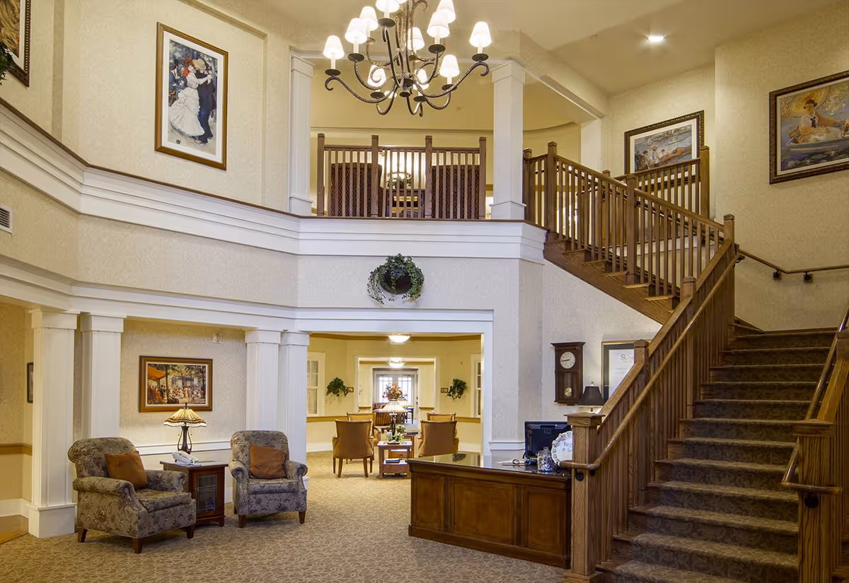 Interior view of a senior living facility lobby with a wooden reception desk, carpeted stairs with wooden railings leading to an upper floor, two upholstered armchairs with a small table and lamp between them, framed paintings on the walls, and a chandelier hanging from the ceiling.