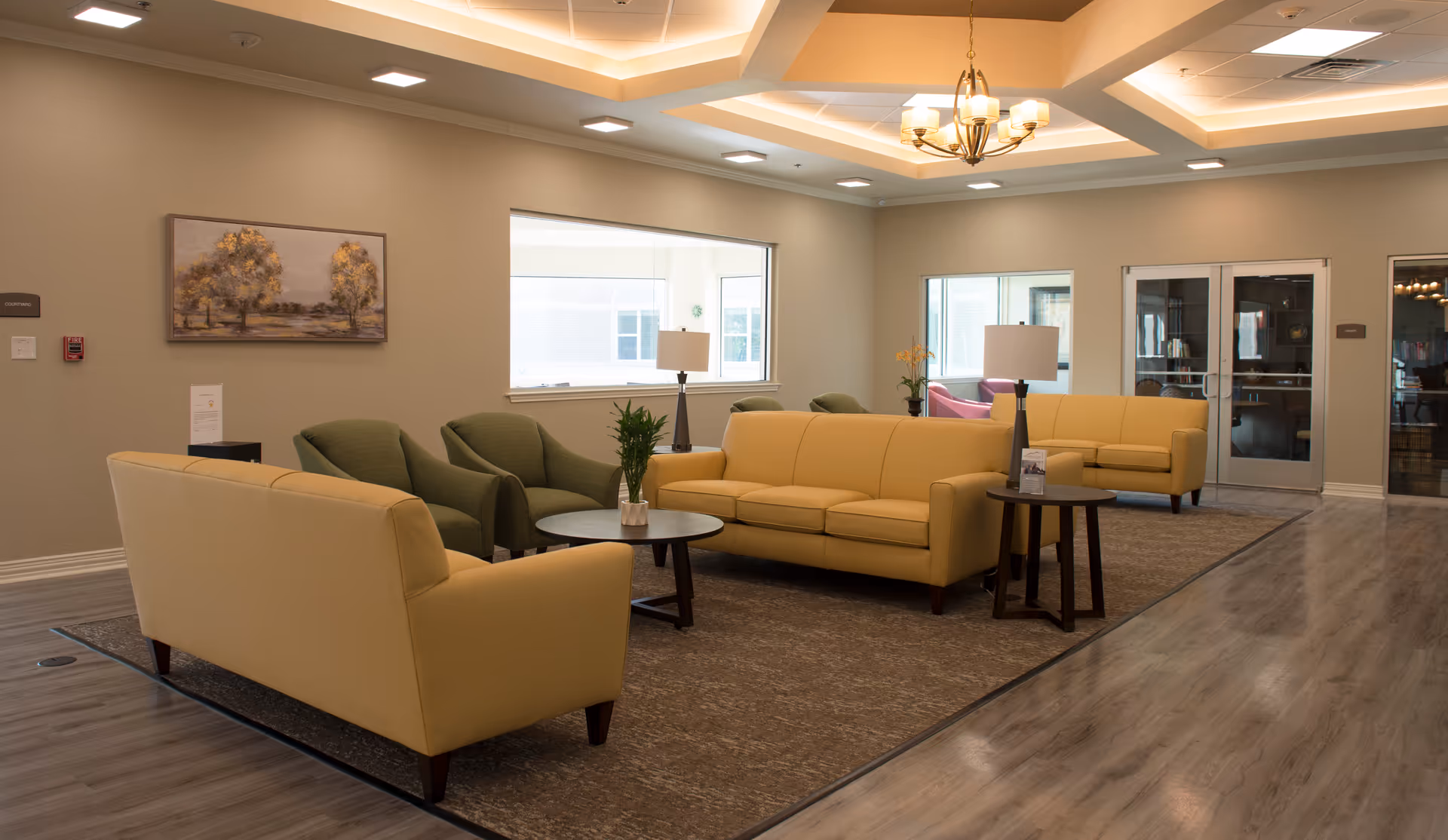 A spacious and well-lit senior living facility lounge area with beige sofas, green armchairs, round wooden coffee tables, and table lamps. The room has a modern ceiling with recessed lighting and a chandelier. There is a large window and glass doors leading to another room, with a painting on the wall and a plant on the coffee table.