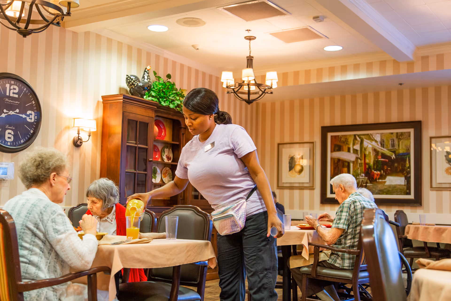 A caregiver in a lavender shirt is pouring orange juice for elderly residents seated at tables in a warmly lit dining room with striped wallpaper, framed artwork, and a wooden cabinet displaying plates.