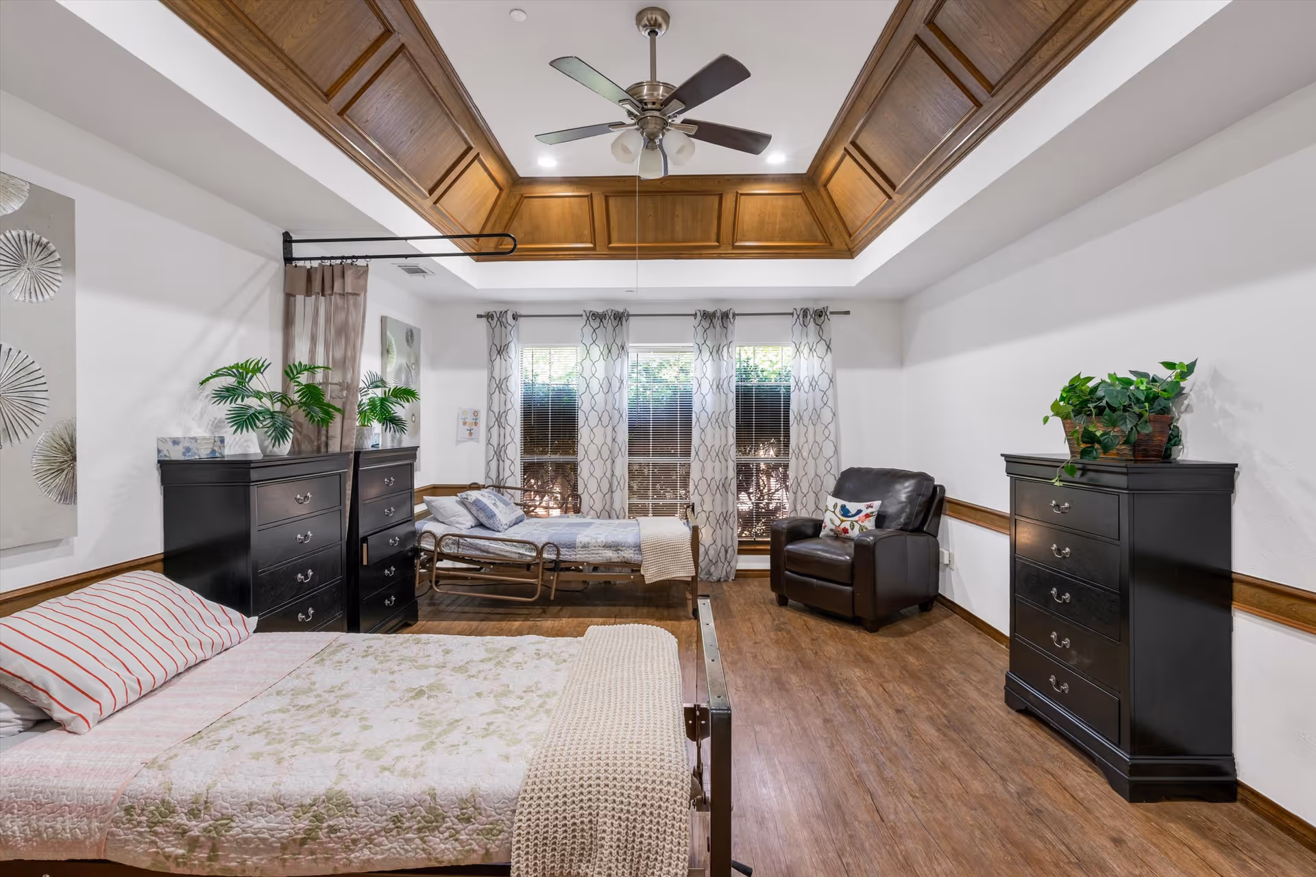 Spacious bedroom with two beds, dark dressers, a leather recliner, and a wood-trimmed coffered ceiling with a ceiling fan.