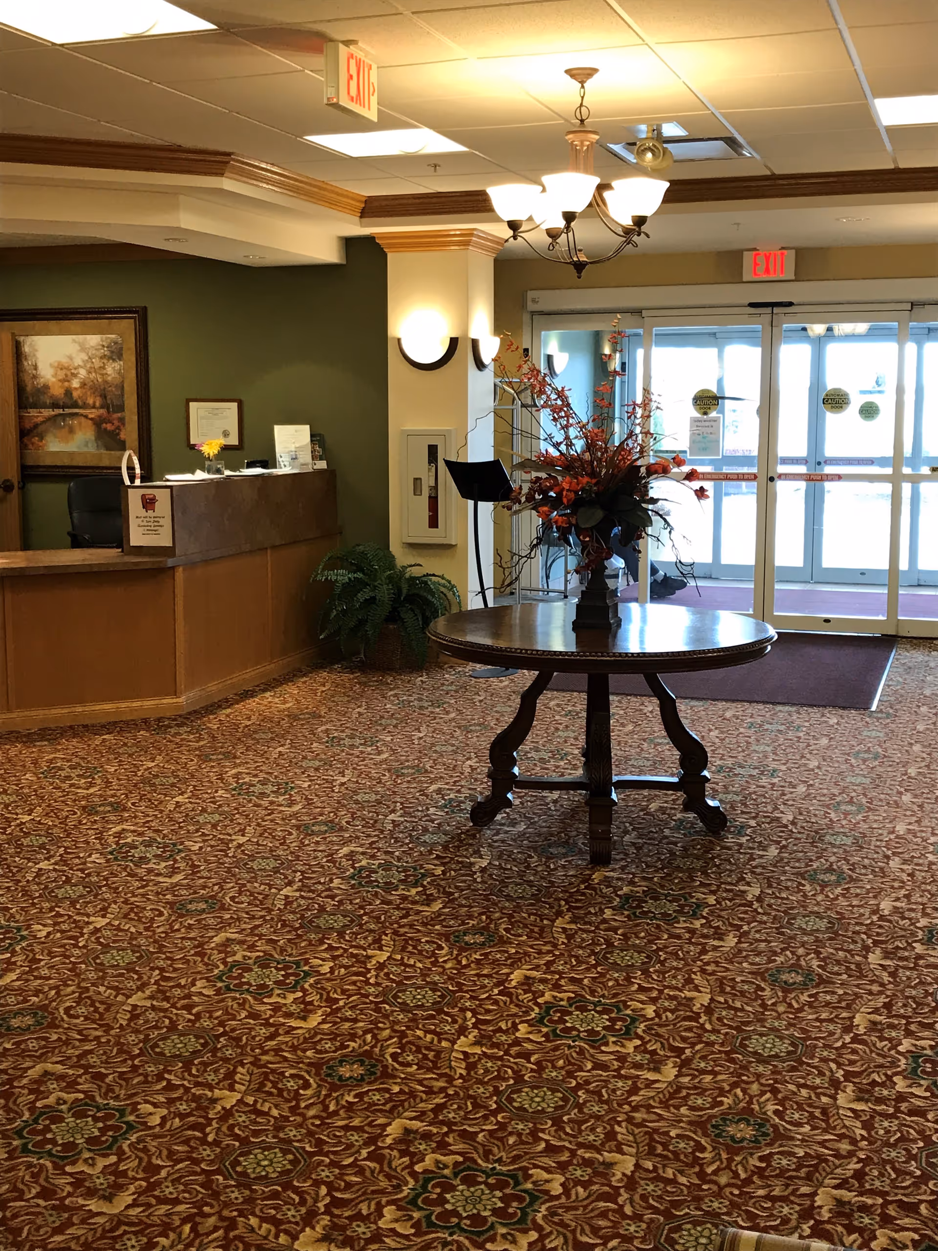 Lobby with a round table holding a floral centerpiece, a reception desk to the left, and glass entrance doors in the background.