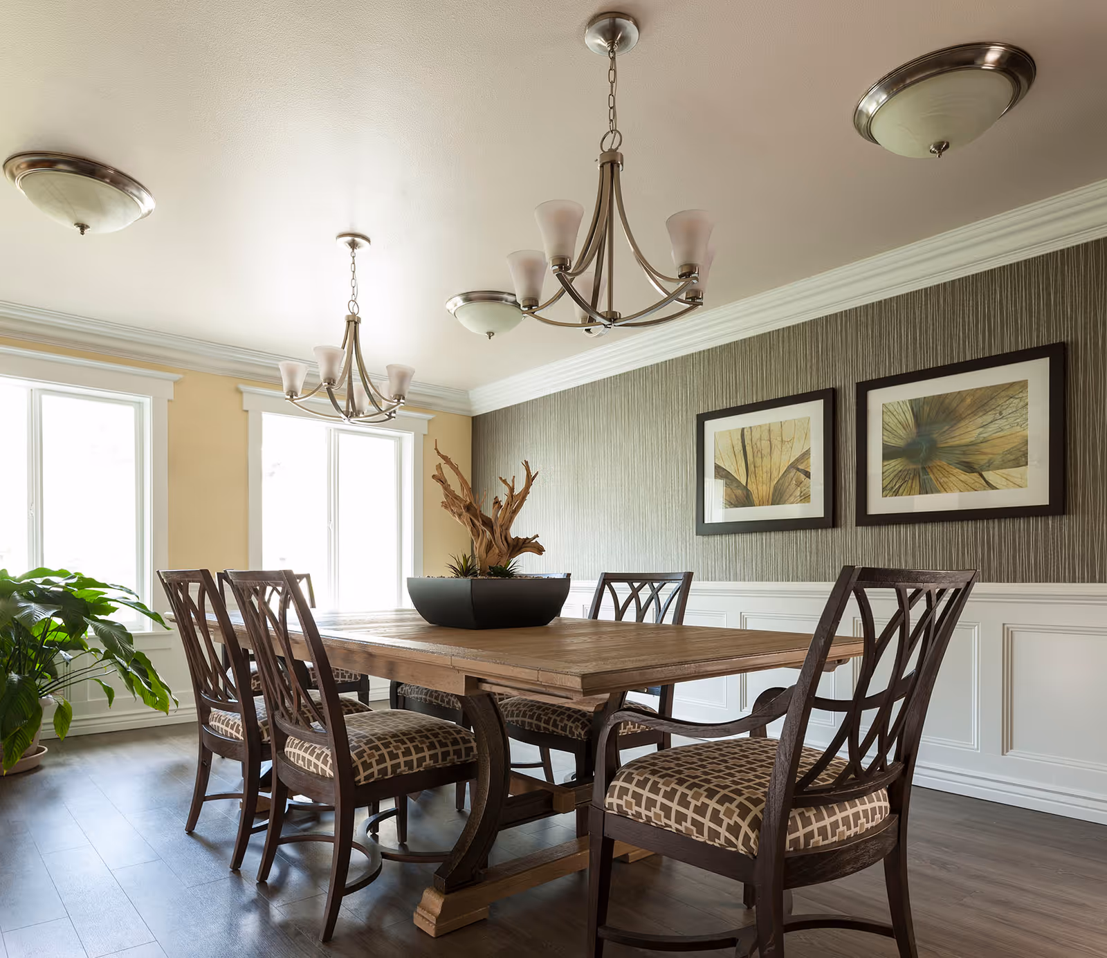 A well-lit dining room with a wooden dining table surrounded by six cushioned chairs with patterned upholstery. The room features two large windows allowing natural light, two modern chandeliers, framed abstract artwork on the wall, and a decorative plant centerpiece on the table. There is also a potted plant near the window.