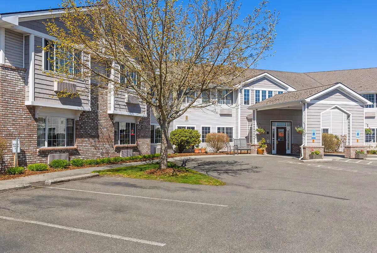 Exterior front entrance of a two-story senior living facility with a tree, landscaped beds, and a parking area.