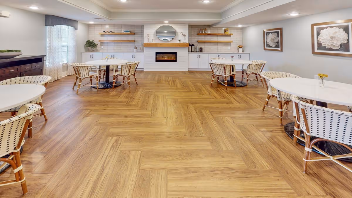 Bright communal dining room with round tables and wicker chairs, wood-patterned floor and a fireplace with shelving along the back wall.