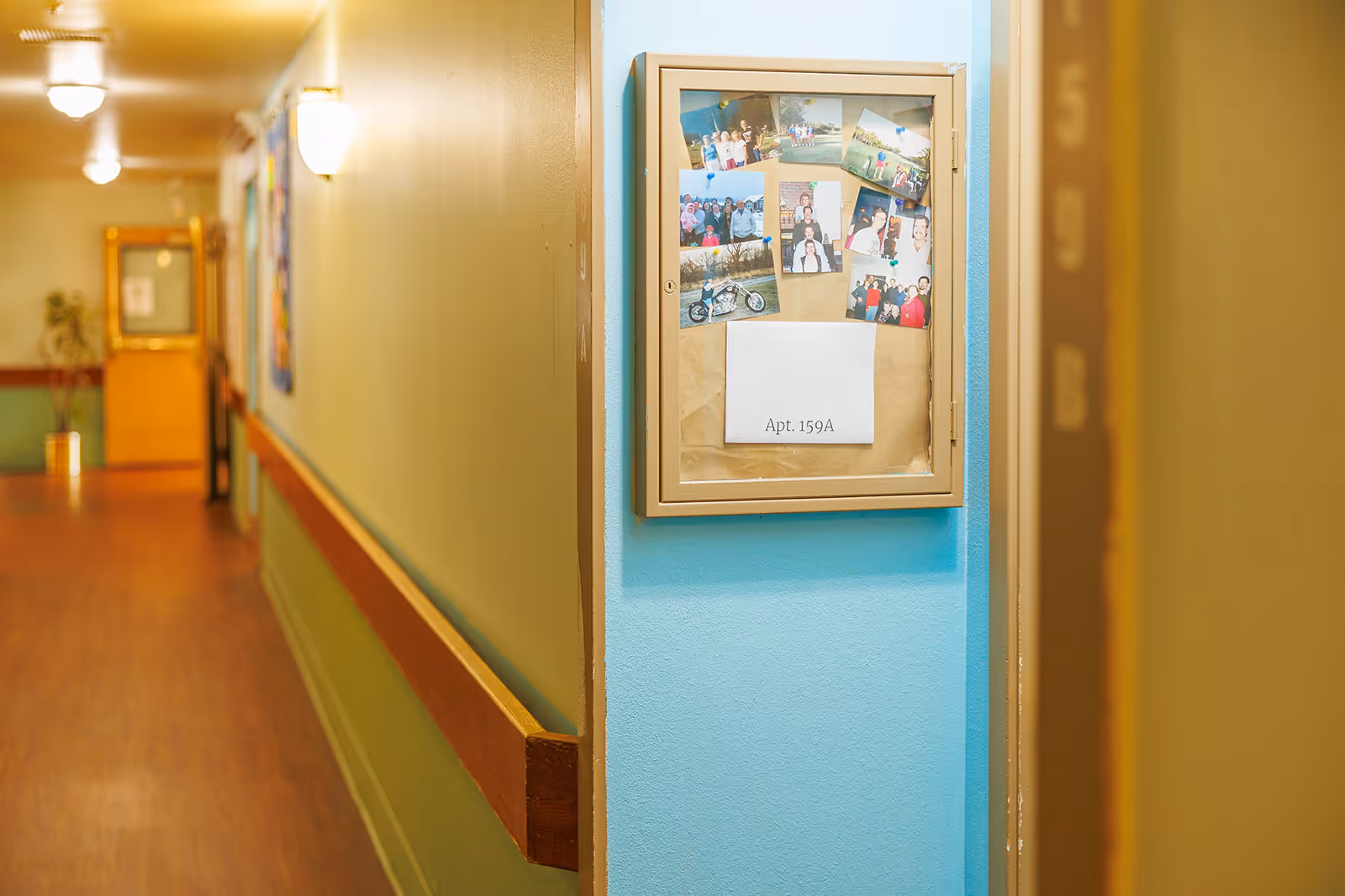A hallway in a senior living facility with wooden handrails along the walls and a bulletin board displaying photos and a sign labeled 'Apt. 159A'. The walls are painted in light green and blue tones, and the floor has a wood-like finish. There are ceiling lights illuminating the corridor.