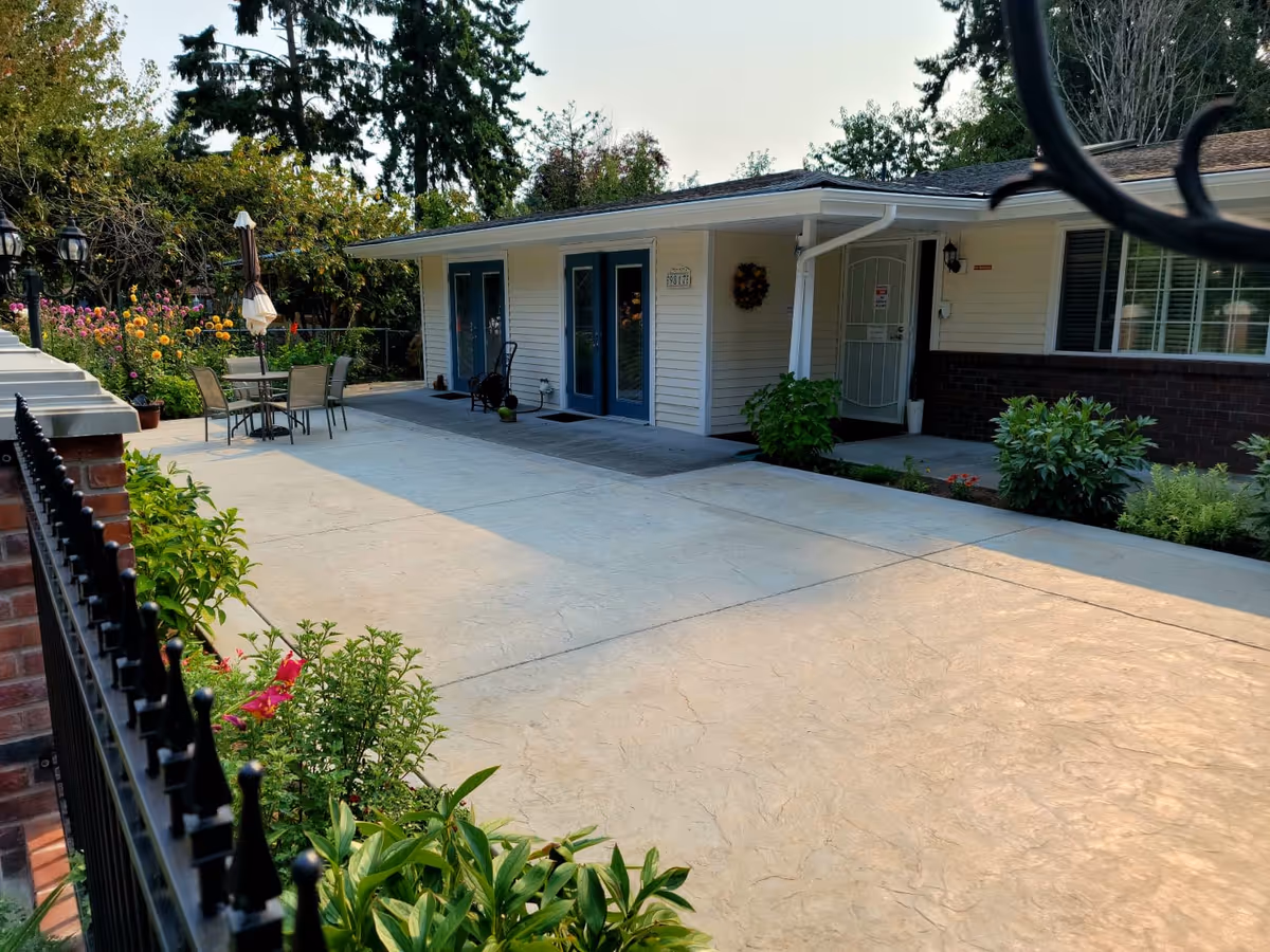 Outdoor patio area of Cambridge Adult Family Home with a concrete walkway, a table with four chairs and an umbrella, surrounded by green plants and flowers. The building exterior is visible with white siding, blue doors, and a covered entrance. Trees and a clear sky are in the background.