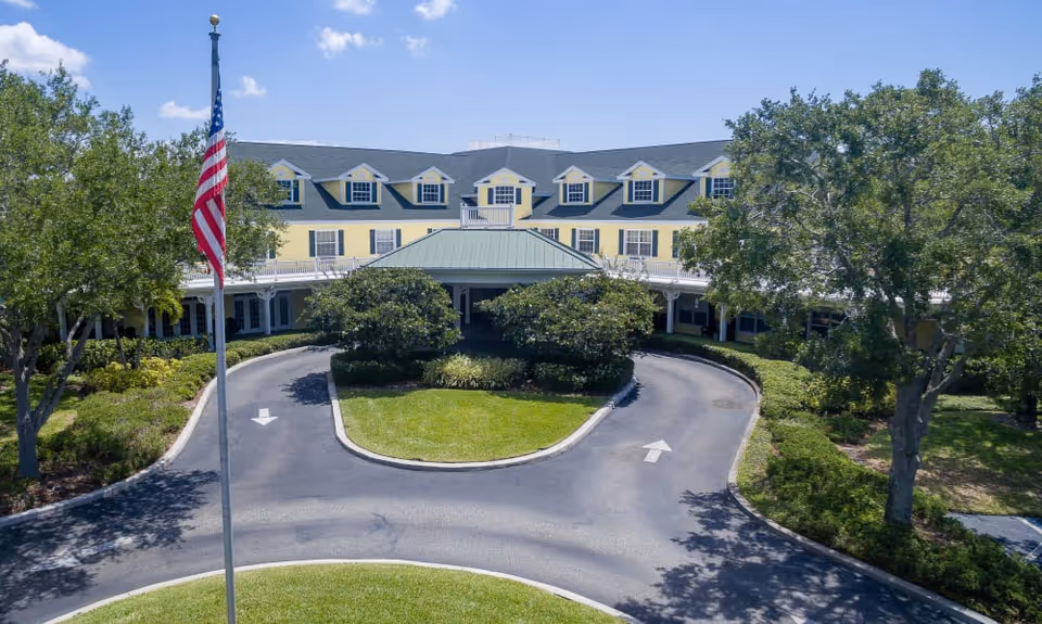 Front exterior view of a large, yellow senior living facility building with multiple dormer windows on the roof, a covered entrance with a green roof, surrounded by trees and landscaping. An American flag on a flagpole is visible in the foreground.