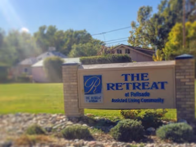 Stone entrance sign reading "The Retreat at Palisade Assisted Living Community" on a lawn with houses and trees in the background.