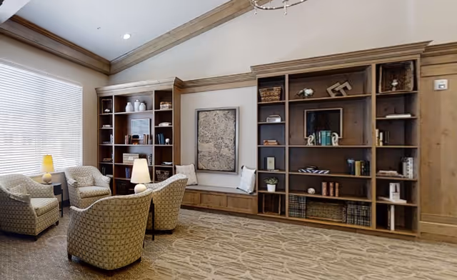 A cozy living room area with four patterned armchairs arranged around a small table with a lamp. Behind the chairs are two large wooden bookshelves filled with books, decorative items, and baskets. A large window with blinds allows natural light to fill the room, and a framed artwork hangs on the wall between the bookshelves. The room has a beige carpet with a subtle pattern and wood trim along the ceiling and walls.
