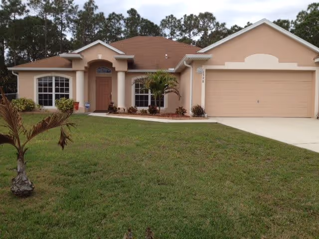 Single-story house with a tan exterior, brown roof, and a two-car garage. The front yard has green grass, small shrubs, and a small palm tree. Trees are visible in the background.