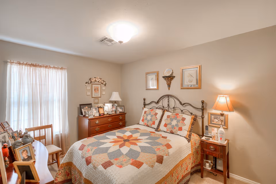 A cozy bedroom with a double bed featuring a colorful quilt and matching pillows. The room has beige walls and a ceiling light fixture. There is a wooden dresser with framed photos and a lamp on it, a small wooden nightstand with a lamp, lotion, and a phone, and a window with sheer curtains letting in natural light. The room is decorated with framed artwork and a decorative wall shelf above the bed.