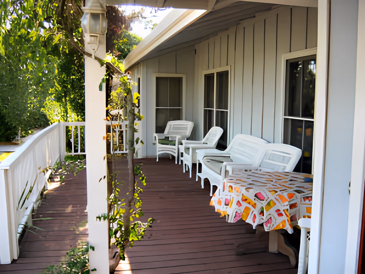 A covered outdoor porch area with white wooden chairs and a bench along the wall of a building. A table with a colorful patterned tablecloth is also present. The porch has a wooden floor and white railing, with greenery visible in the background.