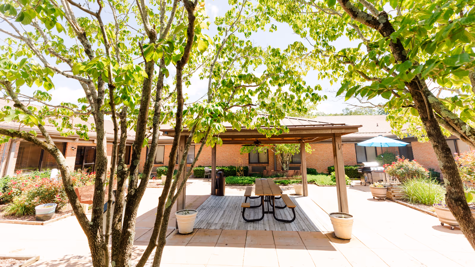 Outdoor courtyard area at Laughlin Healthcare Center with a wooden picnic table under a shaded pergola, surrounded by trees, potted plants, and flowering bushes. The building's brick exterior and windows are visible in the background.
