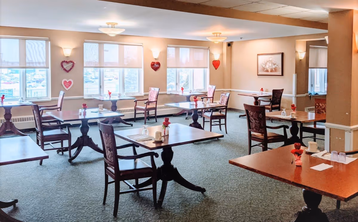 A bright dining room with multiple wooden tables and chairs arranged neatly. Each table has a small red flower centerpiece, cups, and napkins. The room has large windows with white blinds allowing natural light to fill the space. Heart-shaped decorations are hung on the walls near the windows, and a framed floral picture is on one wall.