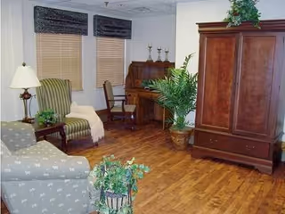 A cozy living room with wooden flooring, featuring a patterned armchair, a striped armchair with a white throw, a wooden side table with a lamp and plant, a wooden desk with a chair, a large wooden armoire, and several potted plants. The room has three windows with blinds and dark valances.