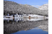 A serene winter landscape featuring a snow-covered mountain and trees reflected in a calm lake, with a few small buildings along the shoreline under a clear blue sky.