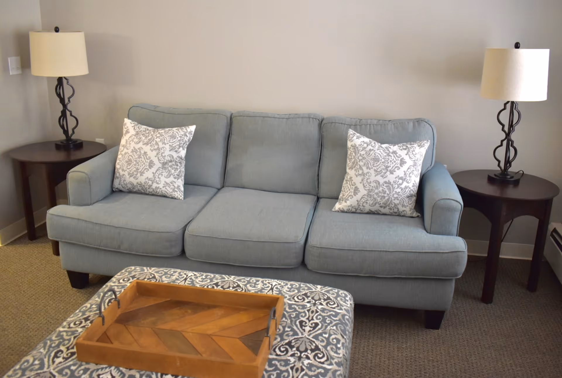 A light blue upholstered sofa with two patterned throw pillows, flanked by two dark wooden side tables each with a decorative lamp. In front of the sofa is a patterned ottoman with a wooden tray on top. The room has beige walls and carpeted flooring.