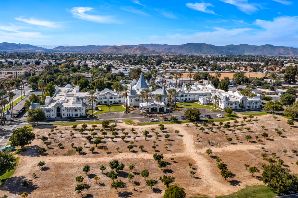 Aerial view of Camelot - A Provincial Senior Living Community, featuring a large white building with castle-like turrets and spires surrounded by green lawns, palm trees, and a parking area. The facility is situated in a suburban area with mountains visible in the background under a blue sky with scattered clouds.