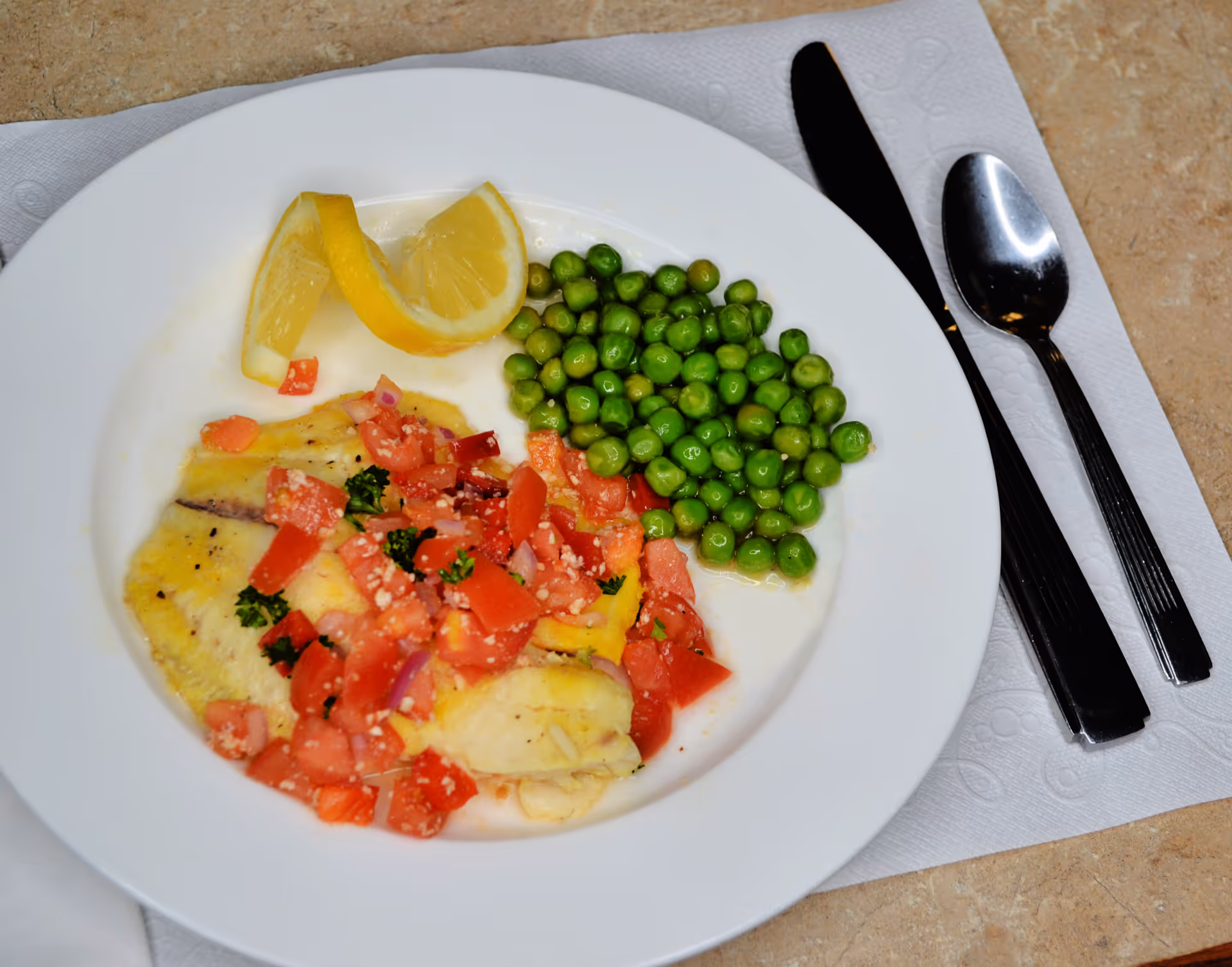 A white plate with cooked green peas, a fillet of fish topped with diced tomatoes and herbs, and two lemon wedges. A knife and spoon are placed on a white napkin to the right of the plate on a beige table surface.