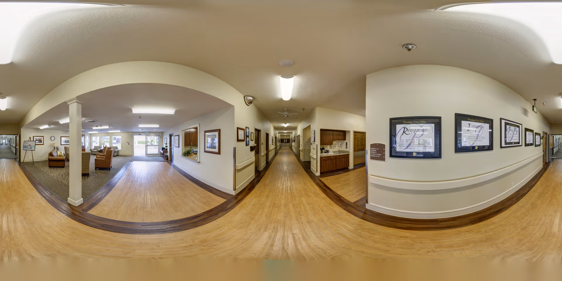 Wide panoramic view of a senior living facility interior hallway with light wood flooring and white walls. On the left side, there is a seating area with chairs and a small table near large windows letting in natural light. The hallway extends straight ahead with framed pictures and certificates on the walls. There is a small kitchenette area with cabinets and a countertop on the right side. The ceiling has recessed lighting and a ceiling fan.