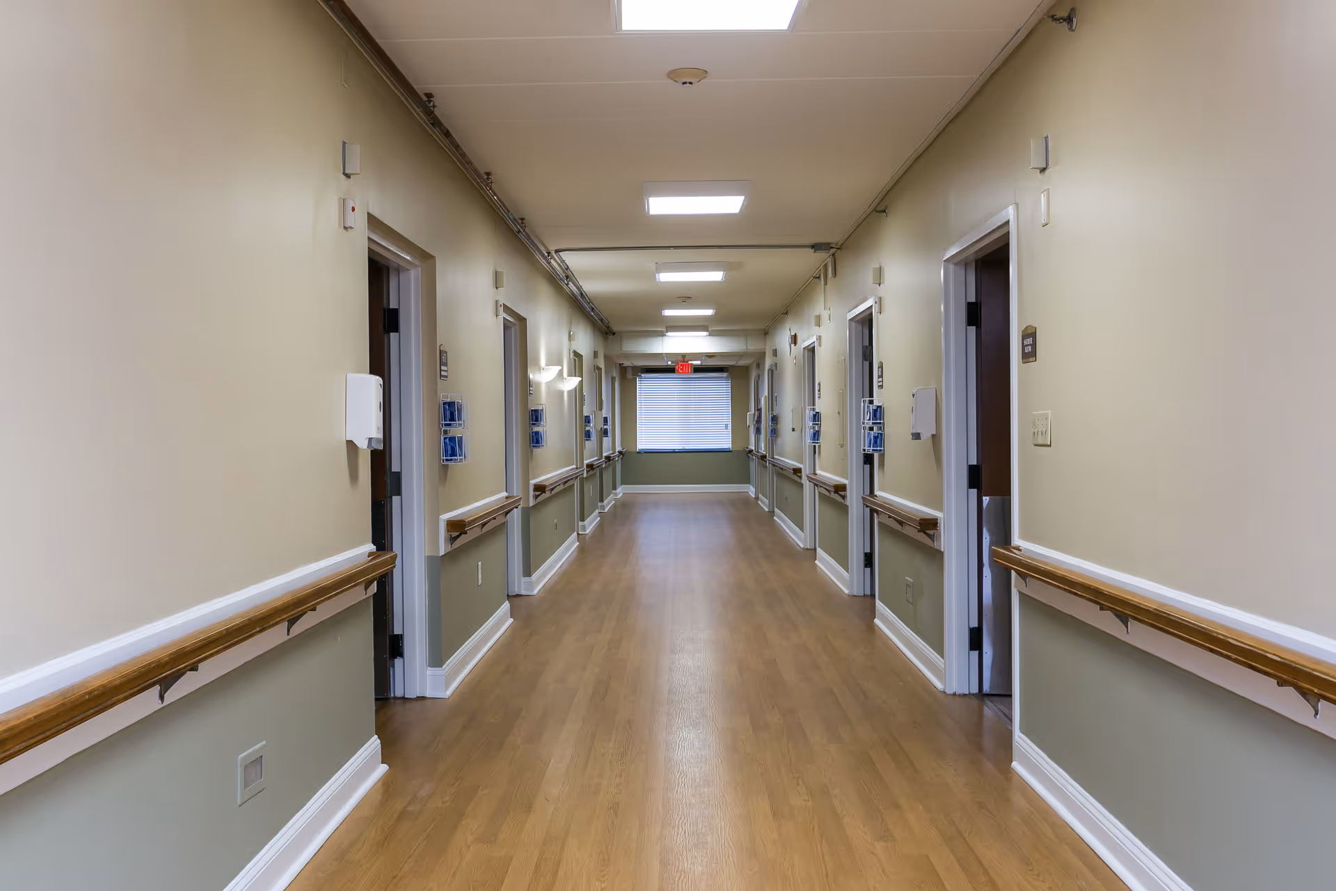 A long, empty hallway in a nursing and rehabilitation facility with wooden handrails on both sides, beige walls, multiple doorways, and ceiling lights leading to a window at the far end.