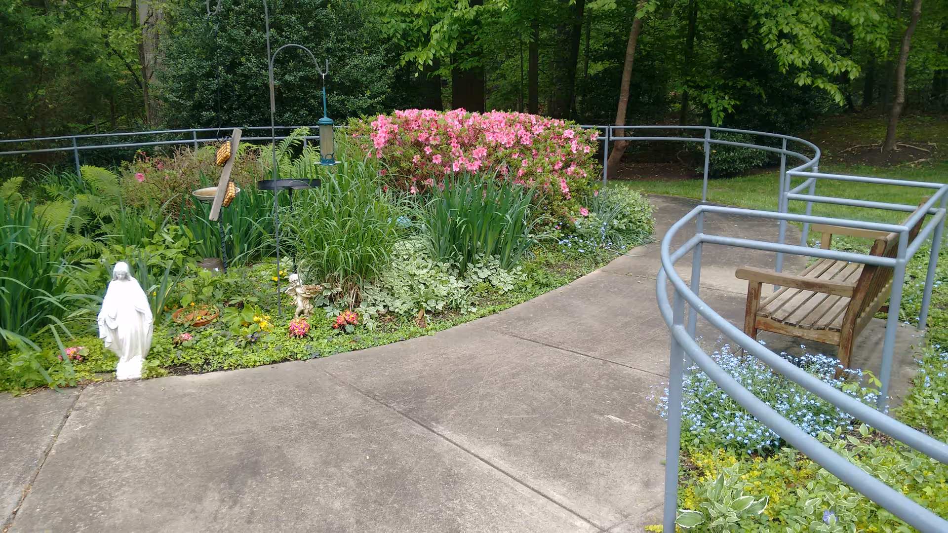 A peaceful outdoor garden area with a curved concrete pathway bordered by metal railings. There is a wooden bench on the right side surrounded by small blue flowers. On the left side, there is a garden bed with various green plants, pink flowers, a white statue, and bird feeders. Trees and greenery are visible in the background.
