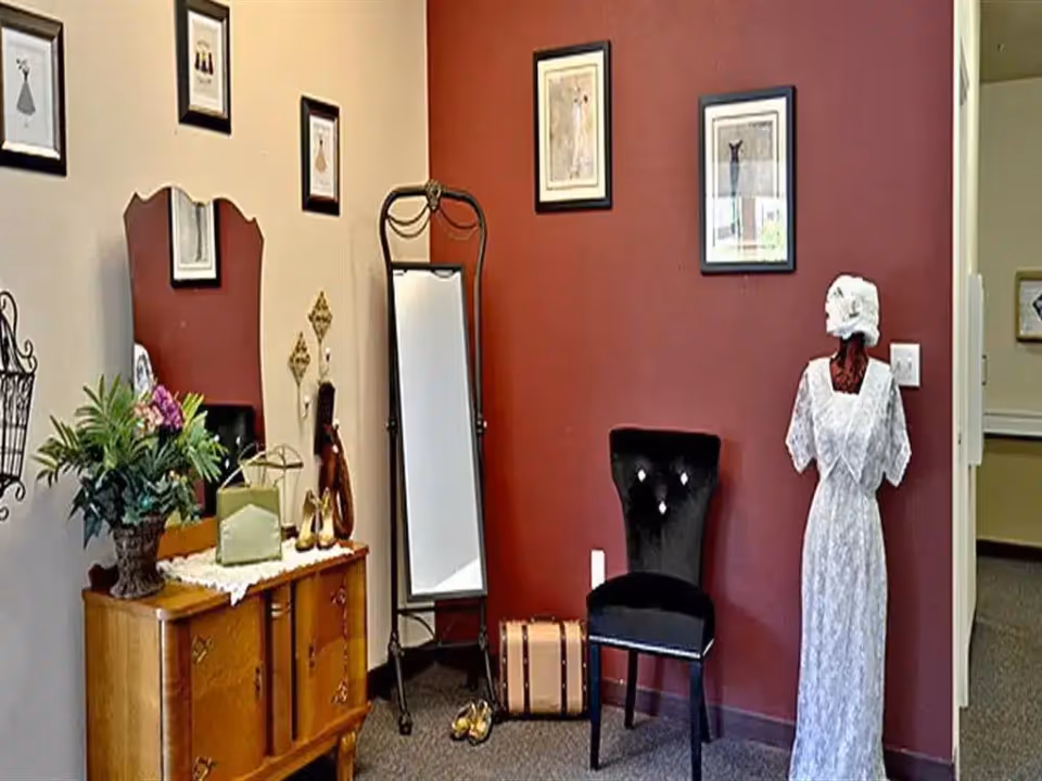 Interior corner decorated with a mannequin in a white dress, a black chair, full-length mirror, wooden dresser with plants and framed pictures on red and beige walls.