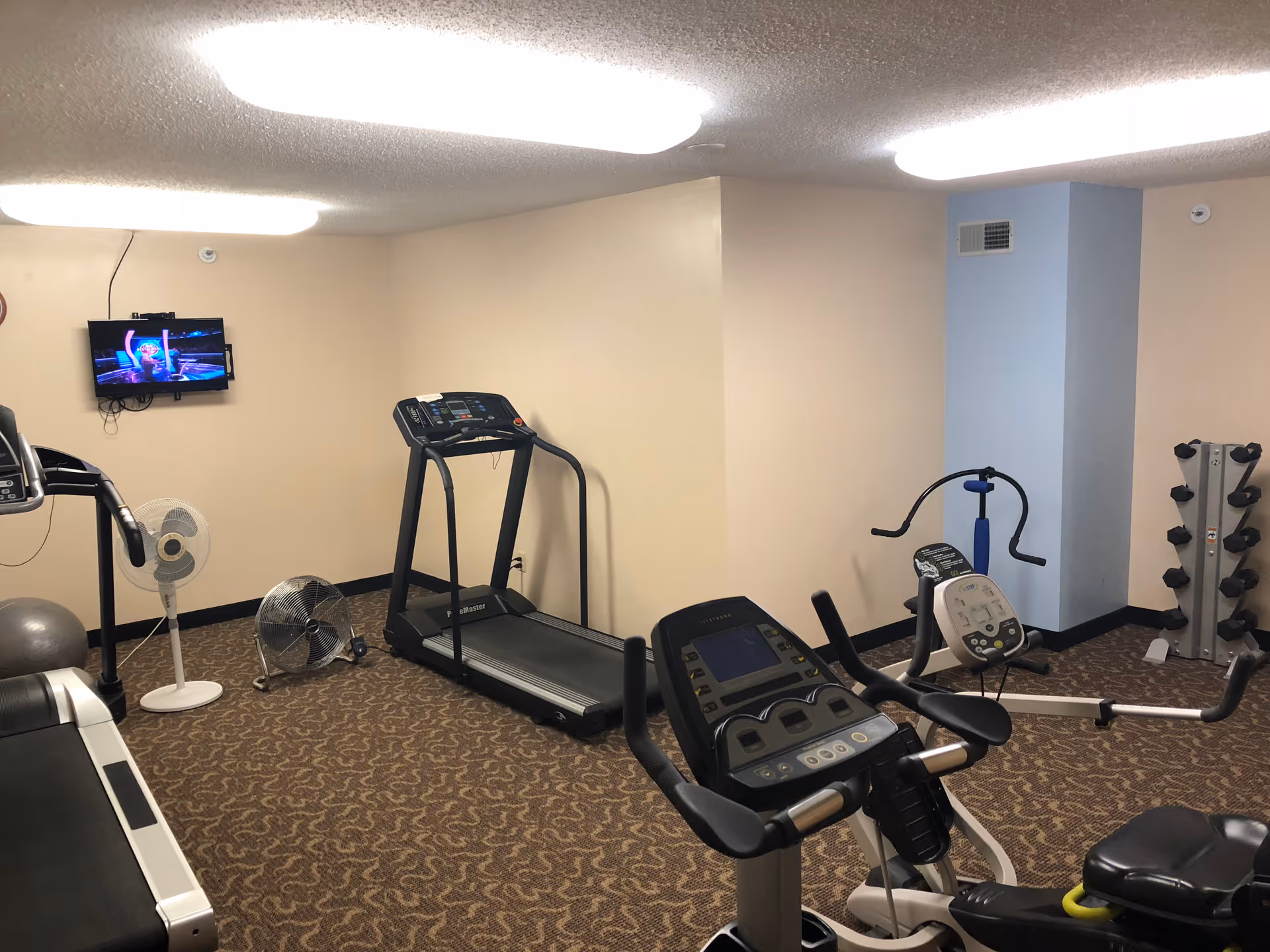 Indoor fitness room with exercise equipment including a treadmill, stationary bike, rowing machine, dumbbell rack, and two fans. A small wall-mounted TV is playing a show. The room has beige walls, a blue accent column, and patterned carpet flooring.