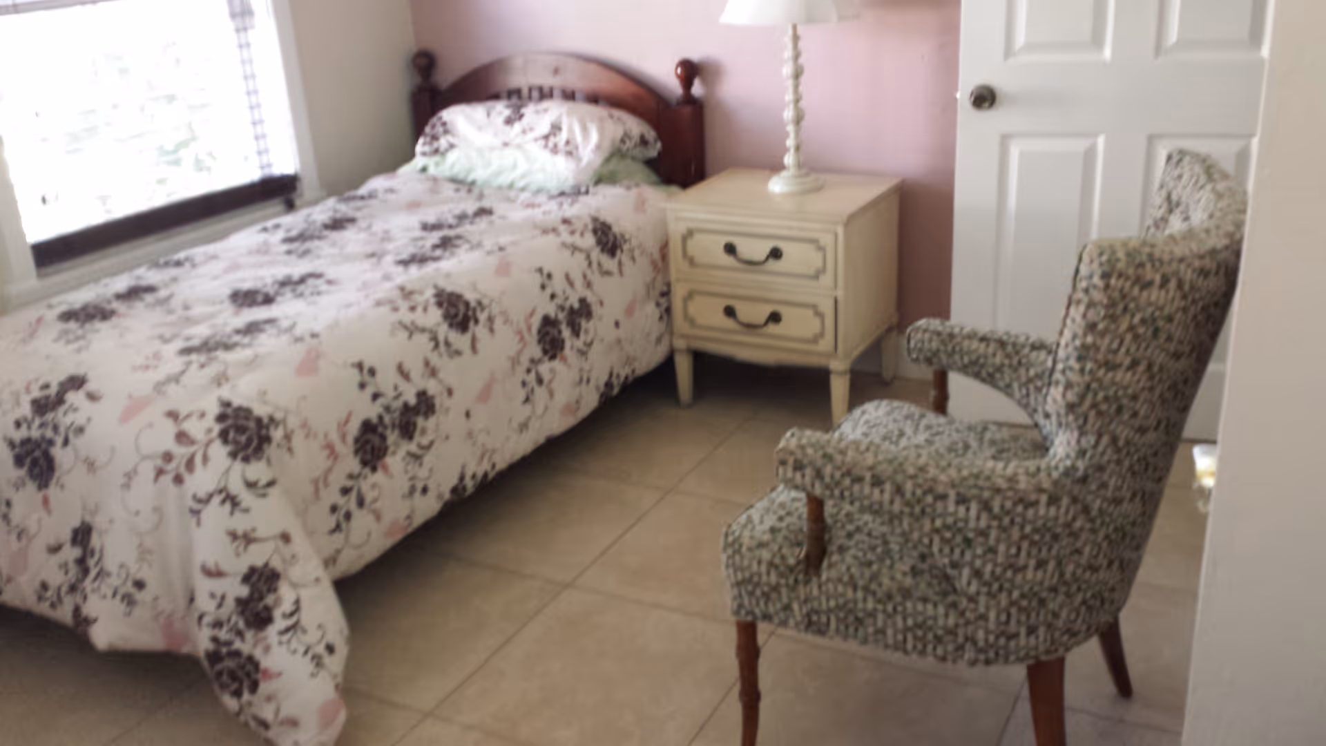 Small bedroom with a twin bed in floral bedding, a nightstand with a lamp, and an upholstered armchair.