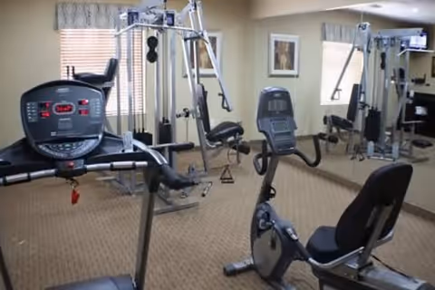 A small fitness room with exercise equipment including a treadmill, a recumbent stationary bike, and a multi-functional weight machine. The room has beige carpet, light-colored walls, a large mirror on one wall, and a window with blinds and a valance.