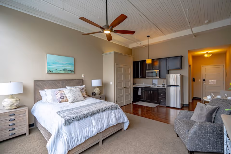 A spacious senior living studio apartment featuring a neatly made bed with decorative pillows, two bedside tables with lamps, a ceiling fan, a small kitchen area with dark cabinets, a microwave, and a stainless steel refrigerator. There is also a comfortable armchair and a small dining table with chairs near the entrance door.