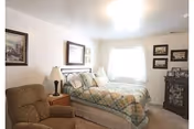 A cozy bedroom with a double bed covered in a plaid quilt, two bedside tables each with a lamp, a comfortable armchair, framed artwork on the walls, and a window letting in natural light.