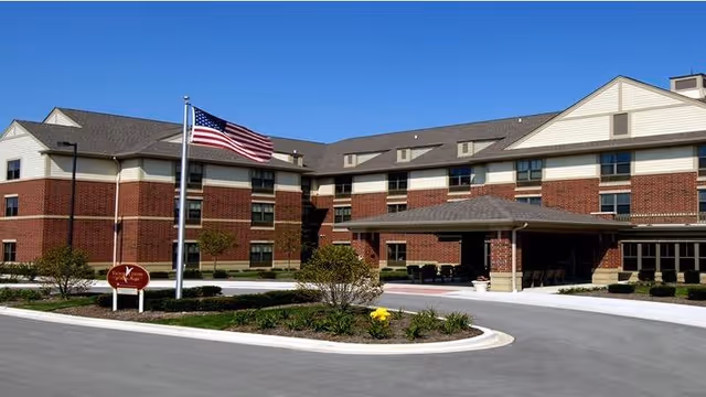 Exterior view of a three-story brick and beige siding senior living facility with an American flag on a flagpole in front and a circular driveway with landscaped greenery and flowers.