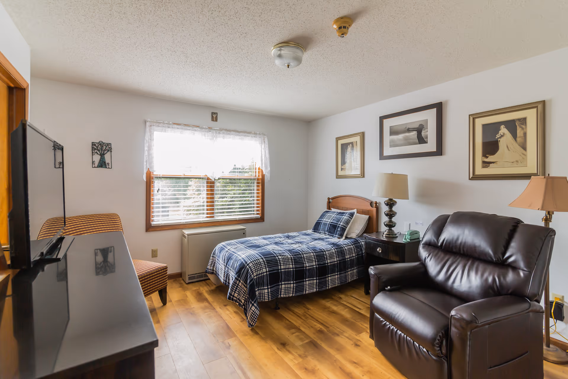 A cozy senior living bedroom with a single bed covered in a blue plaid blanket, a dark brown leather recliner chair, a wooden nightstand with a lamp and a green rotary phone, a window with white lace curtains letting in natural light, framed pictures on the wall, and a flat-screen TV on a dresser.