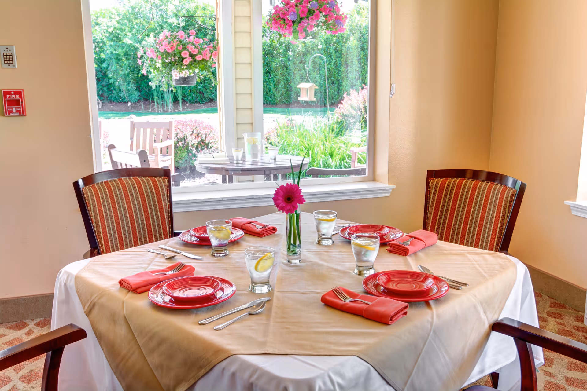 A dining table set for four with red plates, red napkins, and glasses of water with lemon slices. The table has a beige tablecloth with a white undercloth and a single pink flower in a small vase at the center. Behind the table is a large window showing a garden with green plants and pink flowers.