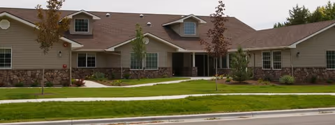 Single-story residential building with beige siding and stone accents, featuring multiple windows and a central entrance with a covered porch. The building is surrounded by a well-maintained lawn with young trees and shrubs, and a sidewalk runs along the front.
