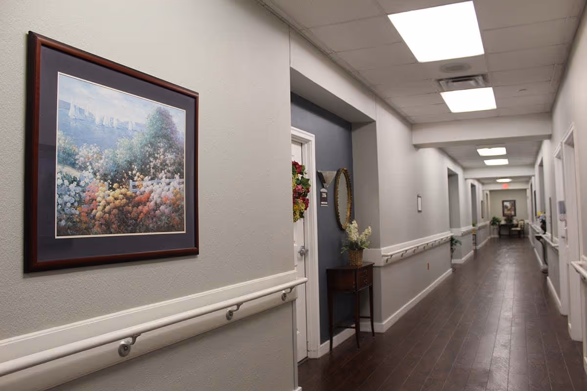 A long, well-lit hallway in a senior living facility with light gray walls, dark wood flooring, handrails on both sides, and several doors along the corridor. There is a framed painting of a colorful garden on the left wall and a small table with flowers and a mirror on the right side. The ceiling has recessed lighting panels.