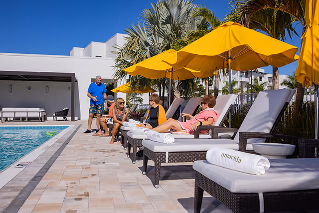 Outdoor pool area at Amavida at Lakes Park with several people relaxing on lounge chairs under yellow umbrellas. The pool is on the left side, and palm trees and buildings are visible in the background.
