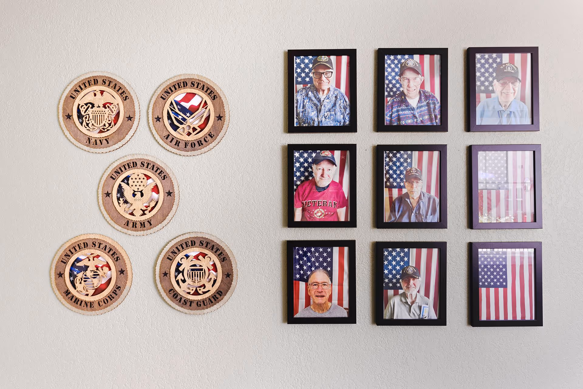 Wall display featuring wooden plaques of United States military branches including Navy, Air Force, Army, Marine Corps, and Coast Guard on the left side. On the right side, there are nine framed photographs of elderly veterans wearing hats and shirts with military insignia, posed in front of American flags.
