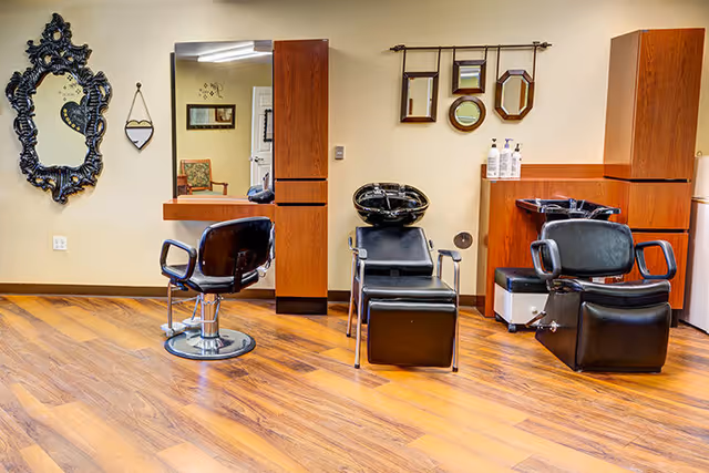 Interior view of a hair salon area with wooden flooring, two black salon chairs, a black hair washing station, a large ornate black-framed mirror on the left wall, and a set of small decorative mirrors on the right wall above wooden cabinets.