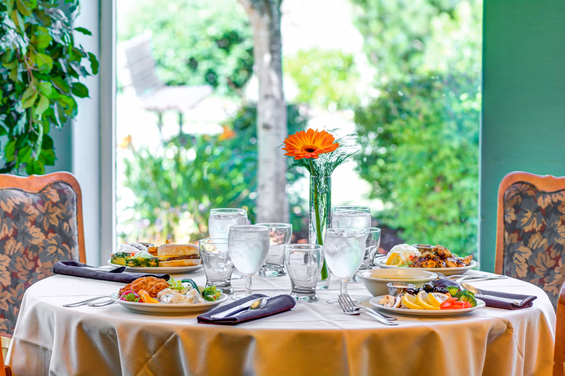 A round dining table set with plates of food, glasses of water, and silverware, with a single orange flower in a vase as a centerpiece. The table is covered with a white tablecloth and surrounded by floral upholstered chairs. A large window behind the table shows a green garden outside.